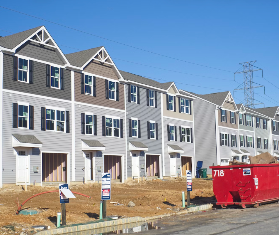 Construction site: 3 story townhomes with garages - grey and beige color schemes. Front doors and dumpster - townhomes have windows on 2nd and 3rd floor - single car garage on first floor.